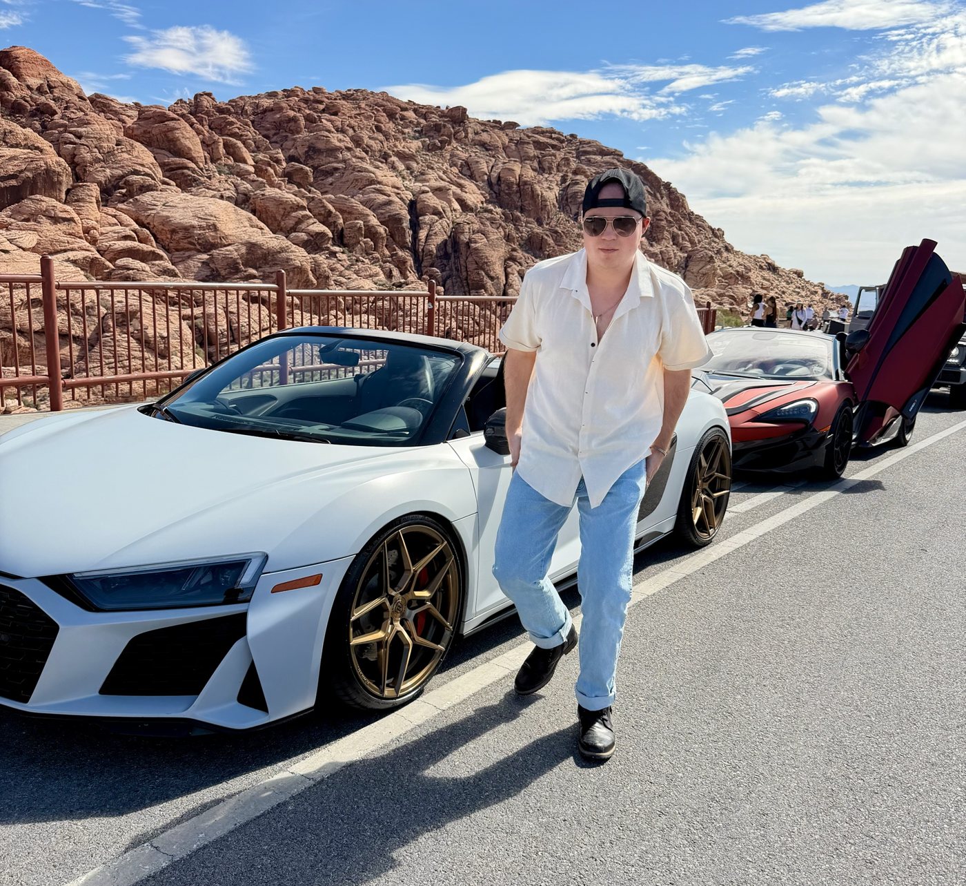 Man in sunglasses and a white shirt beside a white convertible on a desert road lined with red rock