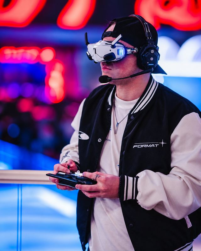 FPV pilot in goggles and headset, wearing a FORMAT varsity jacket, flying with a radio controller under red and blue neon