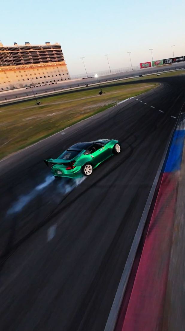 FPV view of a green drift car sliding past a speedway grandstand at sunset