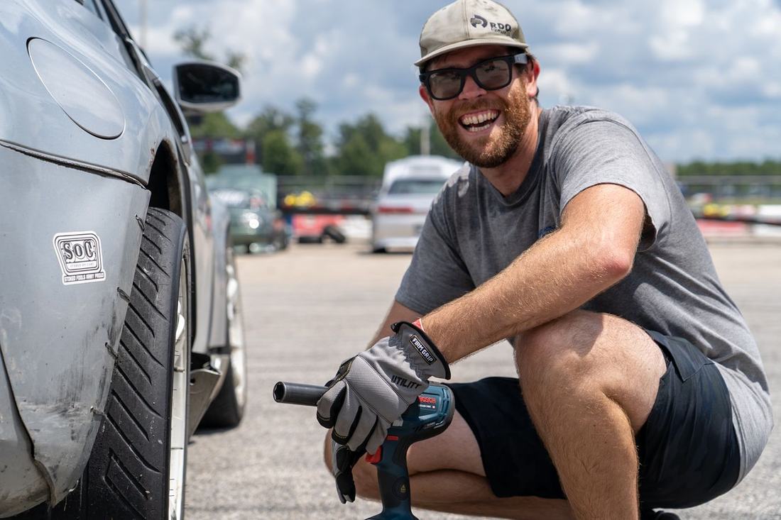 Smiling crew member crouched with an impact wrench beside a car