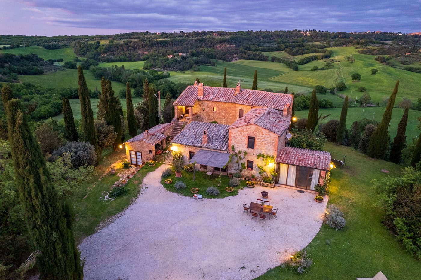 Aerial view of a lit stone farmhouse among cypress trees at dusk